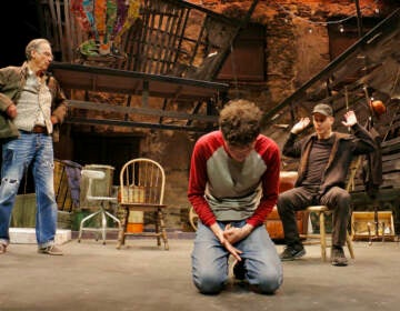 Michael Stahler kneels onstage. Actors David Shriner and Daniel Passer look on behind him.