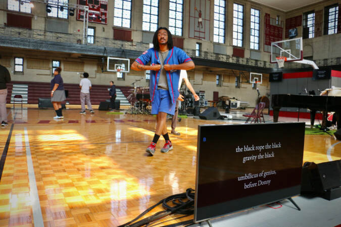 A person wearing a jersey walks around the basketball court as other performers set up instruments.