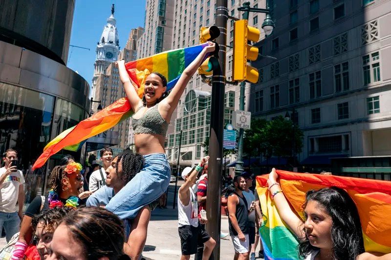 A person holds up a Pride flag in a crowd. City Hall is visible in the background.