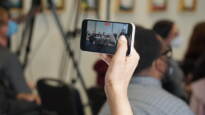 A hand holds up a phone, filming candidates onstage.