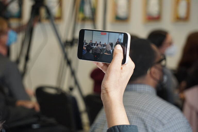 A hand holds up a phone, filming candidates onstage.