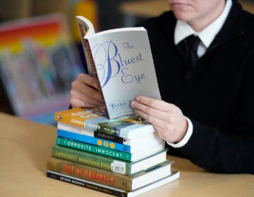 A stack of banned books sits on a table, while someone reads 'The Bluest Eye' by Toni Morrison