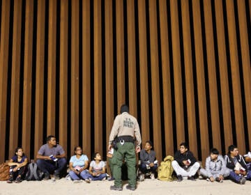 A border patrol agent stands in front of a row of people seated at a wall.