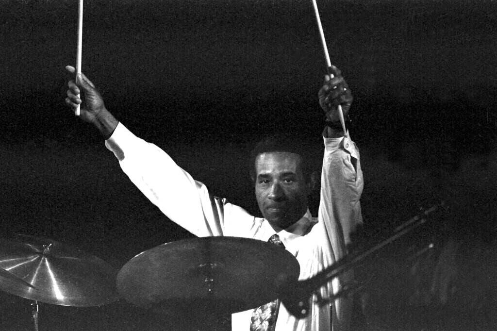 Jazz drummer Max Roach raises his drumsticks in the air as he plays.