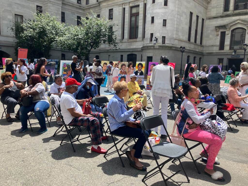 People seated in chairs, walking around outside of City Hall in front of an art exhibition.