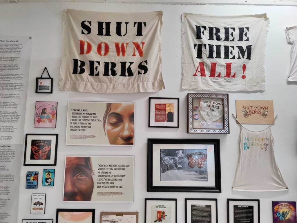 Signs reading "Shut Down Berks" and "Free Them All!" are hung on a wall along with other artwork.