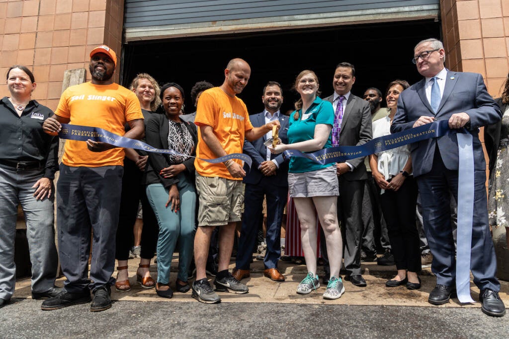 Representatives from Bennett Compost, Philadelphia Parks and Rec, and more gather to cut a ribbon in front of a building.