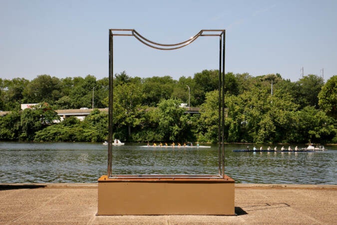 A steel sculpture, behind it are rowers on a river and trees.