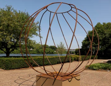 A sculpture made of steel rods forming a sphere is in the foreground. Behind it are trees, the river, and a pathway.