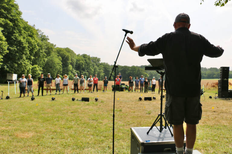 Donald Nally stands in front of a group of people, conducting the choir in an open field.
