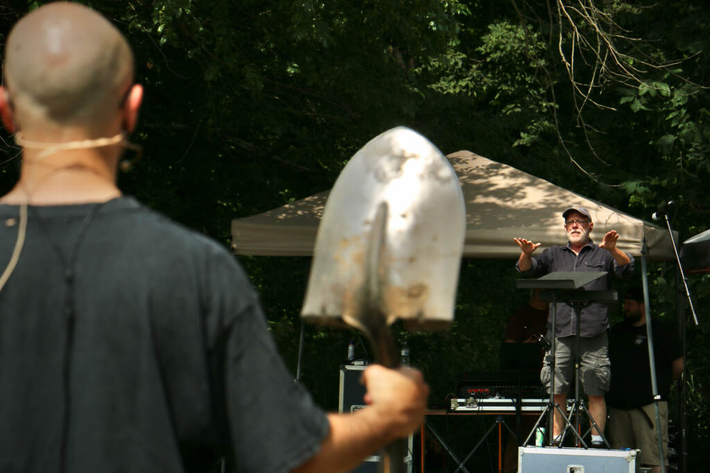 An up-close view of someone holding a shovel in the foreground. In the background Donald Nally conducts.