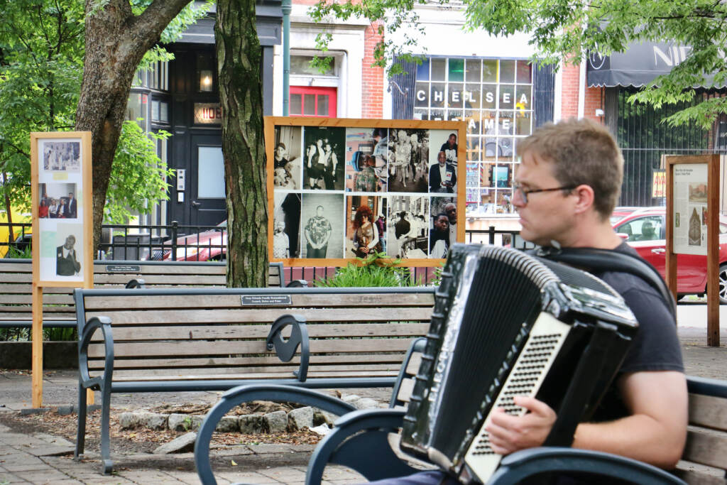 A person plays the accordion in a park with a wall of photos visible in the background.