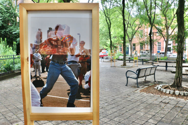 A close-up of a double-exposure photo printed and displayed in a park showing a shirtless man overlayed with a Pride flag.