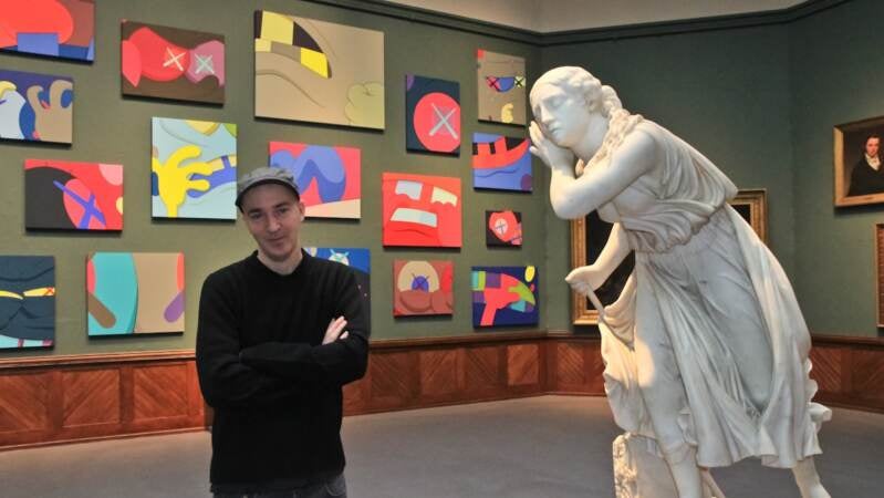 Artist KAWS stands in front of 42 new paintings that focus on the most expressive parts of a cartoon, the hands, eyes and mouth. (Kimberly Paynter/WHYY)