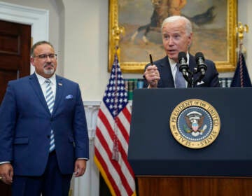 President Joe Biden speaks at a podium. Education Secretary Miguel Cardona listens at left.