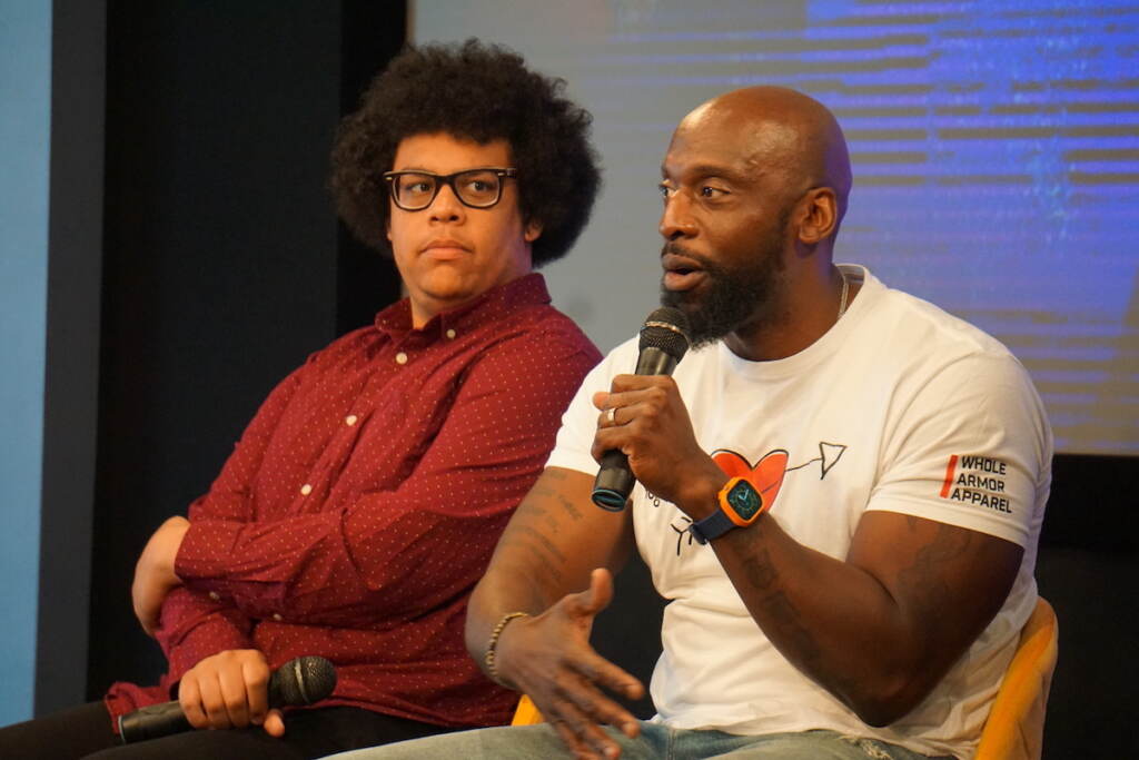 Cory Sharber looks on as Tim Massaquoi speaks into a microphone.