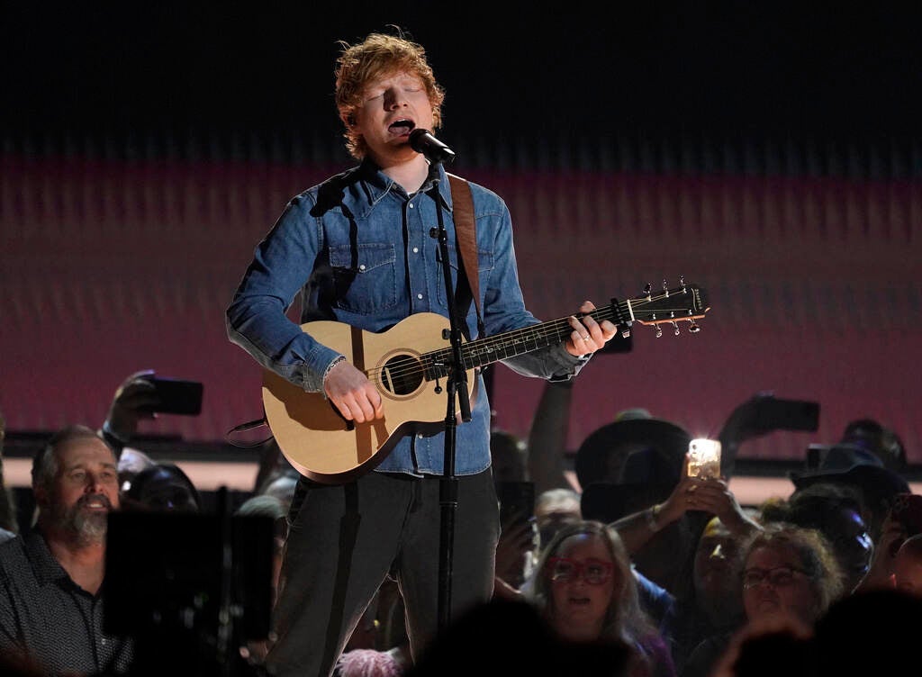 Ed Sheeran sings into a microphone while playing a guitar onstage.