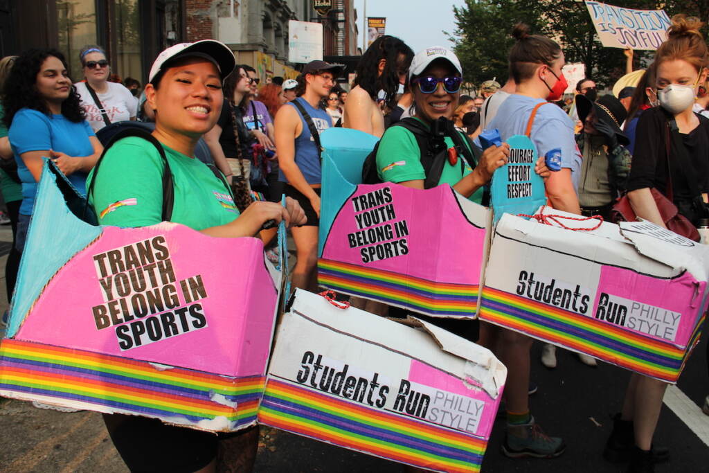 Kim Kim (left) and CC Tellez (right) participate in a protest against Moms for Liberty. Both carry signs that read "Trans Youth Belong in Sports" and "Students Run Philly Style."