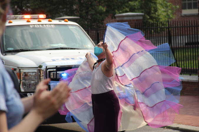 A person with flowing fabric in the colors of the transgender rights flag stands in front of a police car.