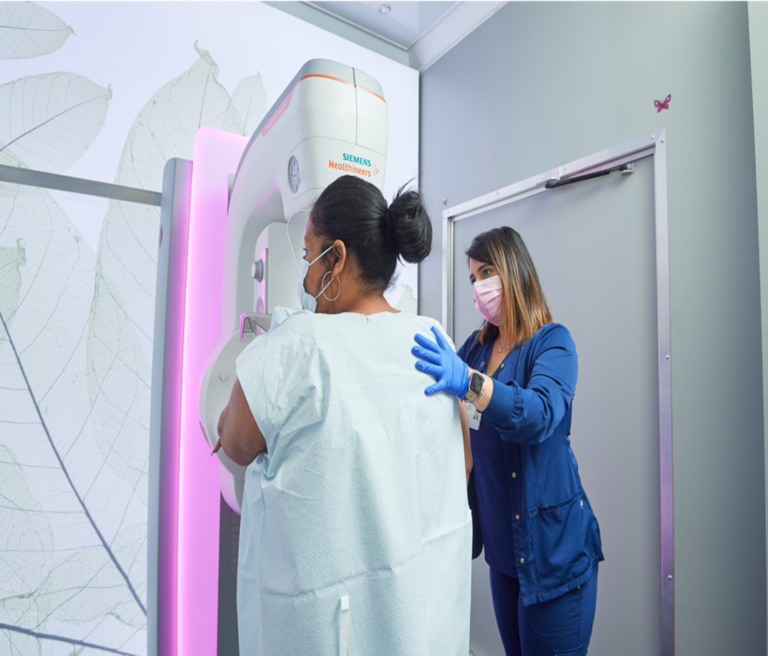 A woman steps towards a mammogram machine, guided by a health care professional.