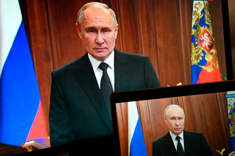 Putin is shown on a screen as he speaks in the background.