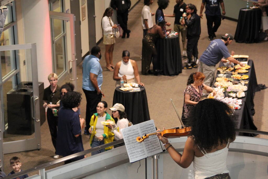 A violinist plays on a staircase landing. Below her people talk, gather around tables, and serve themsevles food.