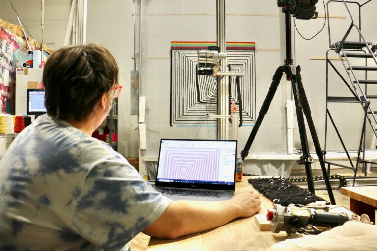 Tim Eads works on a computer. A rug and a tufter machine working on the rug are visible in the background.