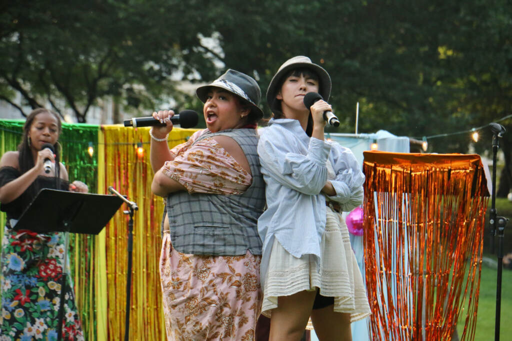 Julia (Vanesa Gomez) and Lucetta (Lexi Thammavong) sing into microphones as they pose back to back.