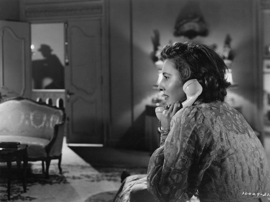 An actress in a black and white film speaks into a telephone. In the background, a shadow of a man is visible.