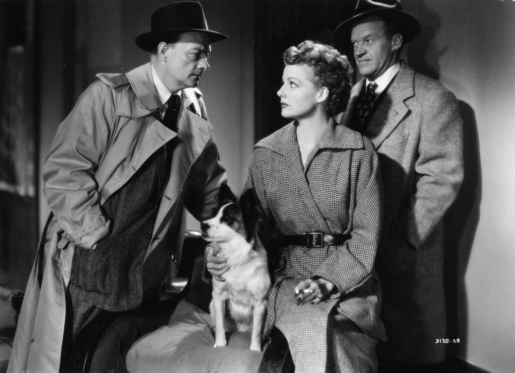 Ann Sheridan sits with a dog on her lap, speaking with a man and another man in the background, in a black-and-white film still from the movie "Woman on the Run"