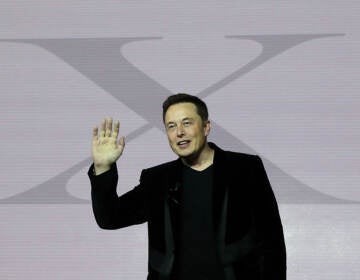 Elon Musk gestures while standing in front of a giant letter 'X'