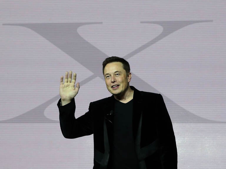 Elon Musk gestures while standing in front of a giant letter 'X'