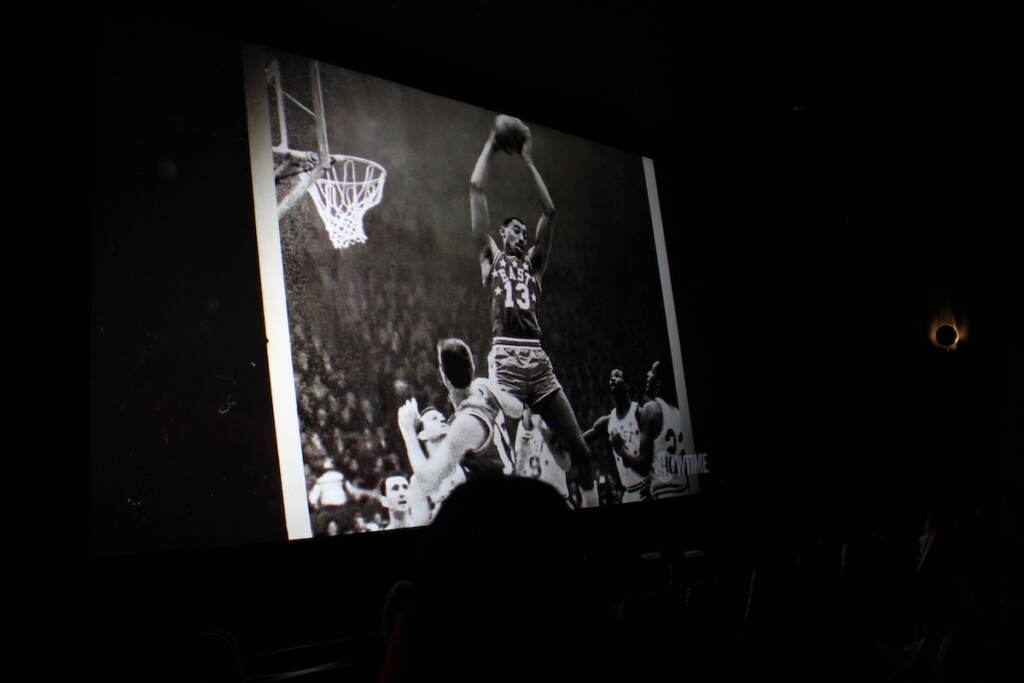 A screen shows a black and white photo of Wilt Chamberlain jumping upwards to shoot the basketball.