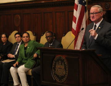 Mayor Jim Kenney speaks into a microphone at a podium at City Hall while other city officials look on.