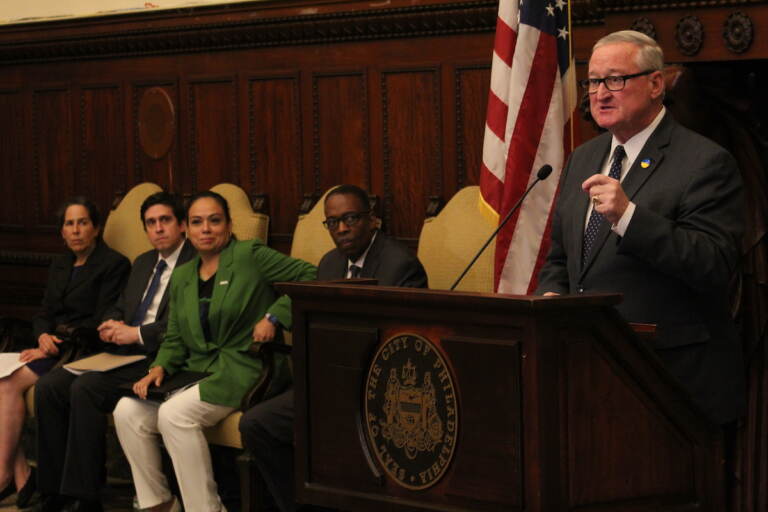 Mayor Jim Kenney speaks into a microphone at a podium at City Hall while other city officials look on.