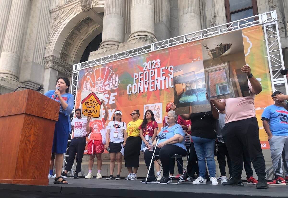 Patty Torres speaks into the microphone. Maria Gracia, seated at the center of the stage, and her family look on.