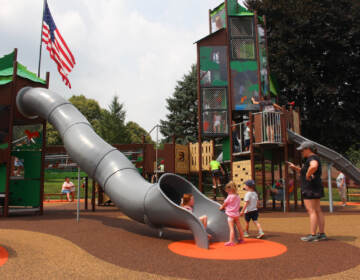 A slide and other structures in the plaground are visible, along with a few children playing, their caregiver, and a U.S. flag waving in the background.