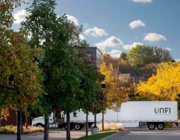 A truck emblazoned with the letters UNFI drives on a road lined with trees.