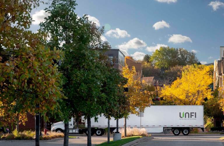 A truck emblazoned with the letters UNFI drives on a road lined with trees.