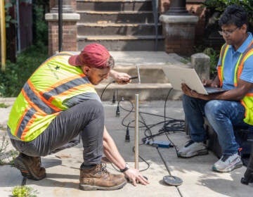 Drexel engineers John DeVitis (left) and K I M Iqbal (right) use a hammer, a rod, a computer, and sensors to record vibrations from the pipe below the sidewalk.