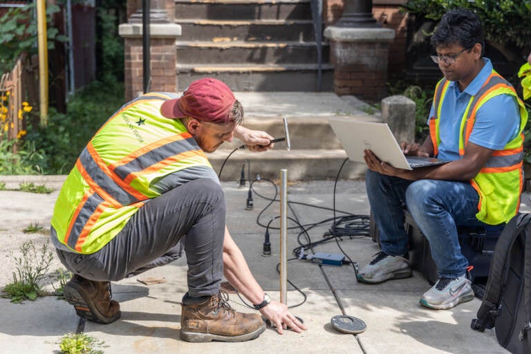 Drexel engineers John DeVitis (left) and K I M Iqbal (right) use a hammer, a rod, a computer, and sensors to record vibrations from the pipe below the sidewalk.