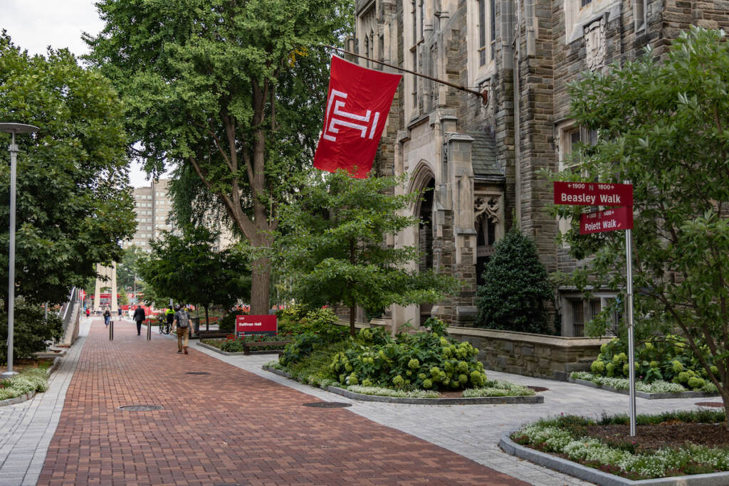 A brick walkway and stone building with a Temple logo flag flying.