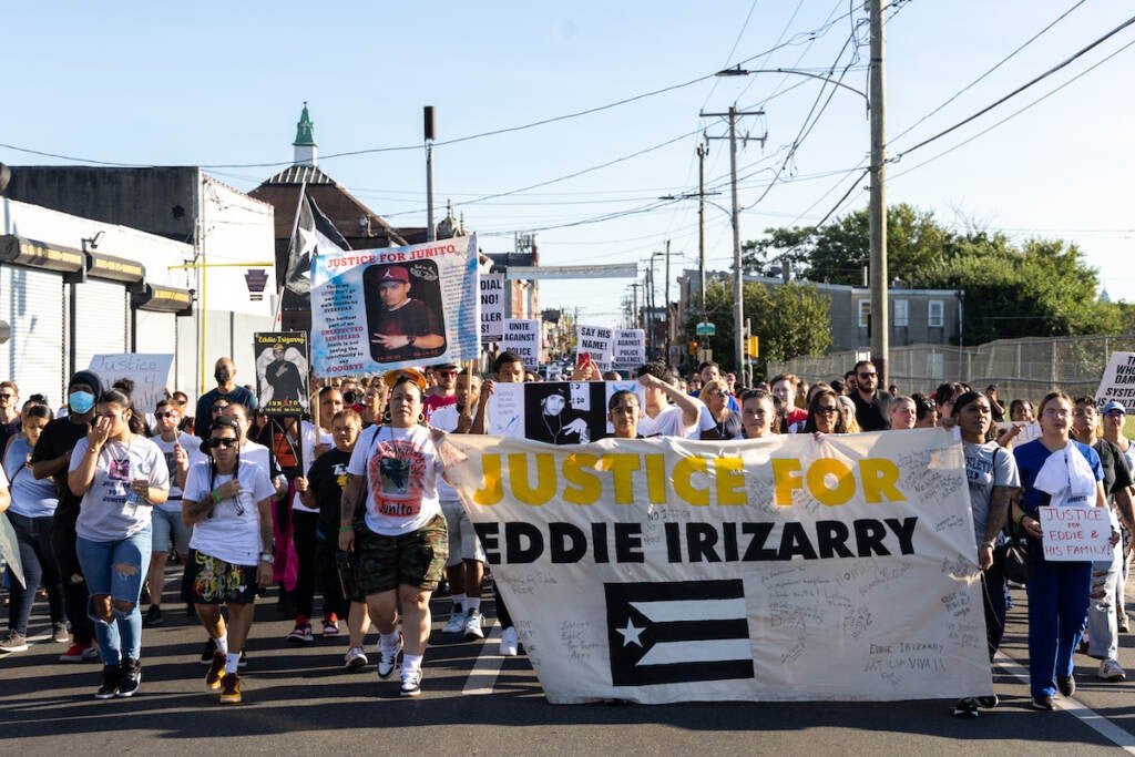 Marchers walk down the street, displaying signs and a large banner that reads "Justice for Eddie Irizarry."