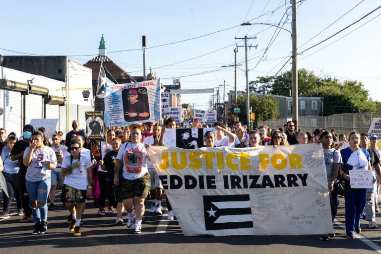 Marchers walk down the street, displaying signs and a large banner that reads 