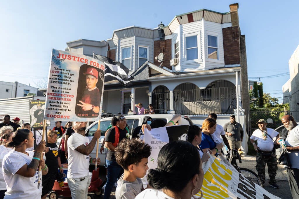 A crowd of people stand together in front of a rowhome, holding signs.