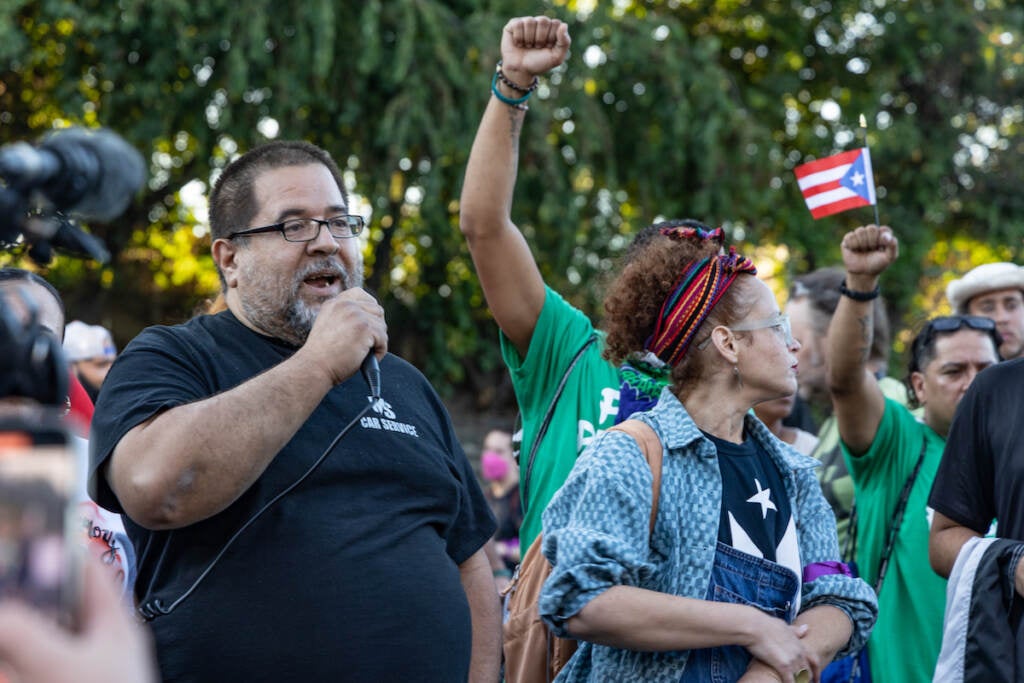 Eddie Irizarry Sr. speaks into a microphone. Marchers stand beside him and around him.