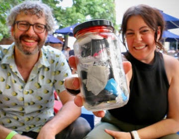 Emily Gallagher holds up a jar of waste as she smiles, seated next to her husband, Austin Elston, who is also smiling.