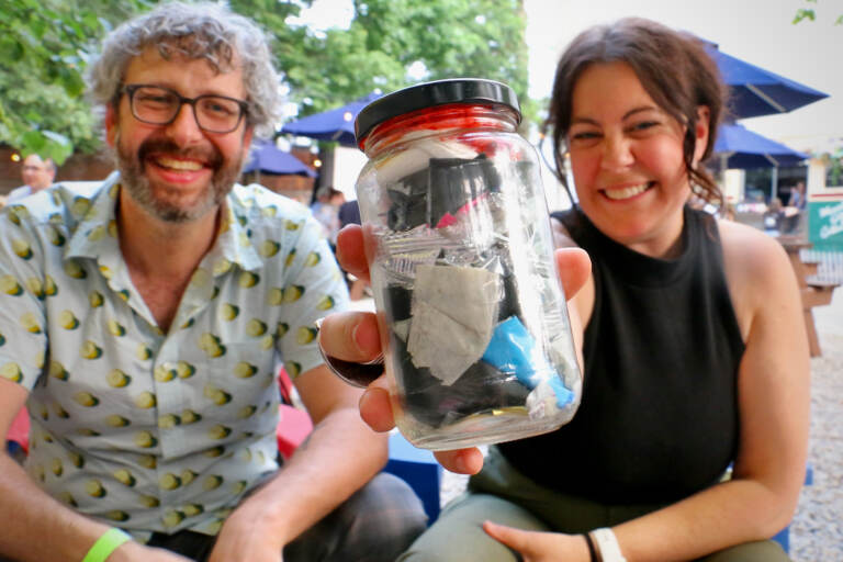 Emily Gallagher holds up a jar of waste as she smiles, seated next to her husband, Austin Elston, who is also smiling.