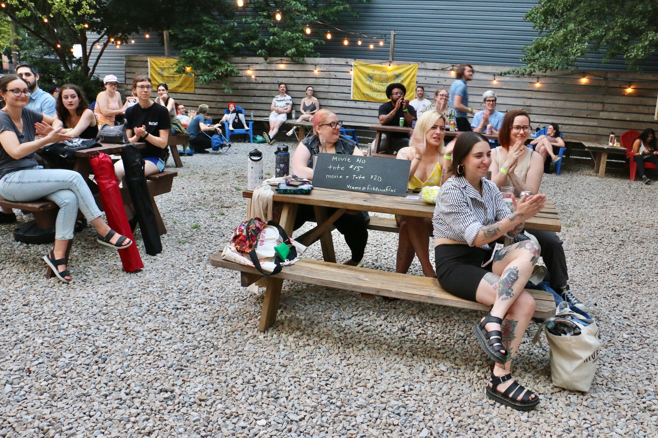 A crowd of people seated at picnic tables outdoors applaud.