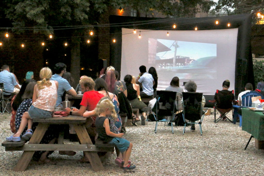 A crowd of people seated outdoors watch a movie being shown on a projector screen.